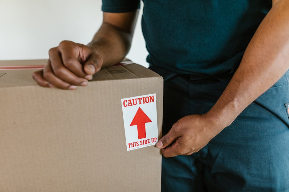 Two women standing amidst a collection of packed cardboard boxes in an indoor space with a textured grey wall background. One woman, dressed casually in beige trousers and a checked shirt, holds a med