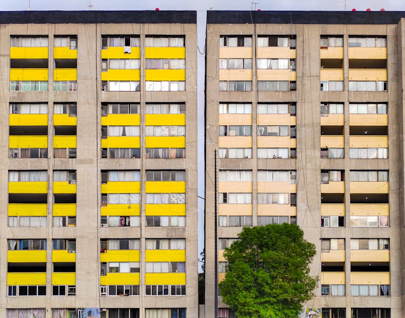 Photograph of a tall, multi-storey residential building in Hornsey with a façade featuring numerous small, uniformly spaced windows. The building is surrounded by mature trees with green foliage, som