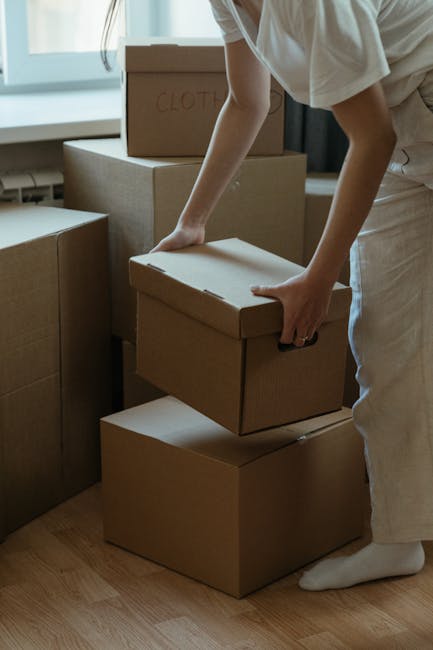 A person wearing white clothing is stacking and organizing cardboard boxes in an indoor space, likely during a home relocation process. The individual is holding a small rectangular box, preparing it for transportation, with several larger boxes around them, some labeled with permanent marker, such as 'CLOTH.' The room has natural light coming through a window, illuminating the boxes placed on a wooden floor. The person appears to be in the process of packing or unpacking, with their hands positioned on a box that is being either placed down or picked up. This scene depicts the packing and handling phase of moving, which is part of the professional furniture transport and home removals services provided by Man With a Van Hornsey. The environment is orderly, and the boxes are secured with tape, ready for loading into a van for transportation, highlighting the logistical aspects of a local move near Hornsey Station, HORNSEY.