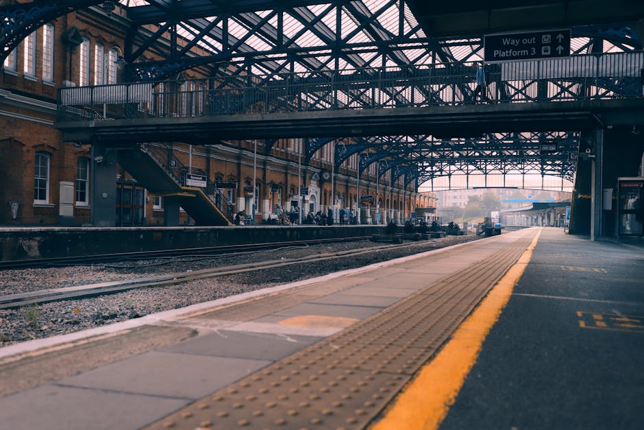 The image depicts a train station platform with a covered metal and glass canopy overhead, supported by steel beams and trusses, creating an industrial aesthetic. The platform surface is paved with concrete and features tactile paving strips near the edge for safety, along with painted yellow lines indicating the boundary for train safety zones. To the left, there are brick station buildings with large windows, and a staircase with yellow safety markings leading up to a footbridge that spans across the platform, allowing passengers to access different sides. Several people are visible standing or sitting along the platform, some waiting, while others are engaged in activities. Railway tracks run parallel to the platform, with gravel beds and sleepers visible between the rails, indicating active train routes. In the background, a train is arriving or departing, and the station environment is well-lit by natural light, suggesting daytime. This scene illustrates the typical setting for a transportation hub, pertinent to house removals, logistics, and transport services provided by companies like Man With a Van Hornsey during home relocation and furniture transport activities.