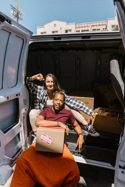 Two women are preparing for a home relocation by loading cardboard boxes into the back of a large silver van parked outdoors under a clear sky. One woman, wearing a plaid shirt, is sitting inside the 