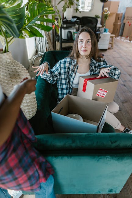 Two women are preparing for a home relocation by loading cardboard boxes into the back of a large silver van parked outdoors under a clear sky. One woman, wearing a plaid shirt, is sitting inside the 